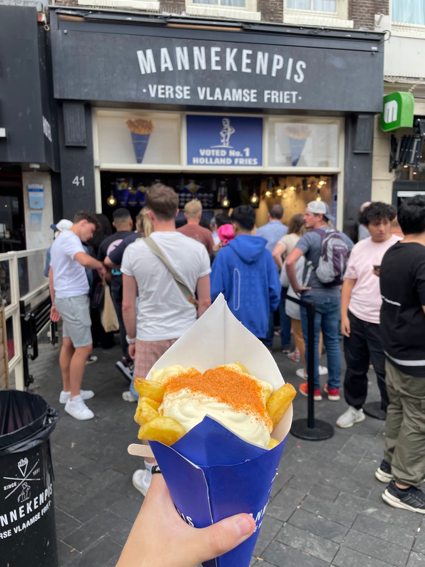 A paper cone of thick-cut frites topped with mayo and spice powder, held up in front of the Mannekenpis frites shop in Amsterdam